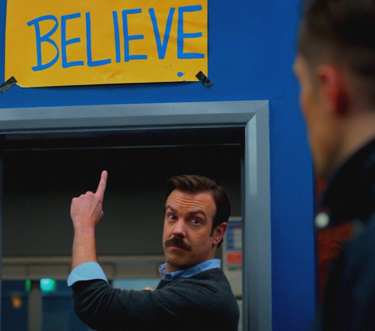 Ted Lasso pointing at a sign reading ‘Believe’
