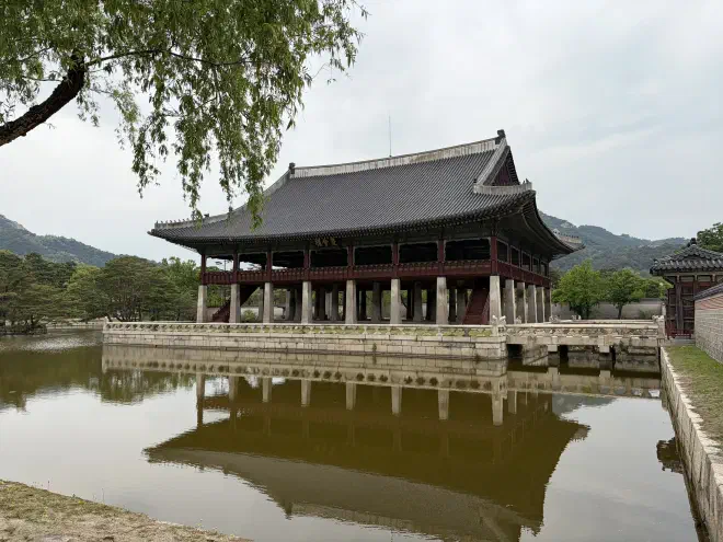 A large open-sided traditional Korean building sits on a stone plinth in the middle of a lake. A bridge sits on its right joining it to land