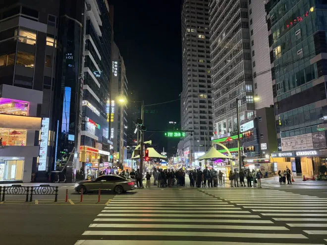 A wide pedestrian crossing with people waiting and tall buildings in the background