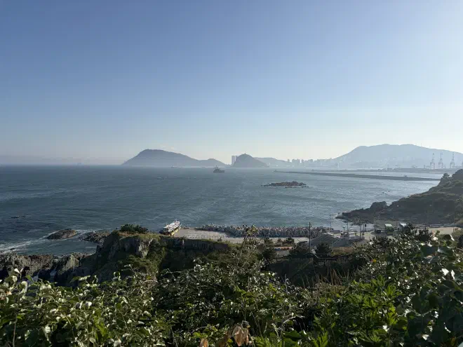 An ocean with a city and mountains in the distance and a concrete dock with a boat docked at it in the foreground