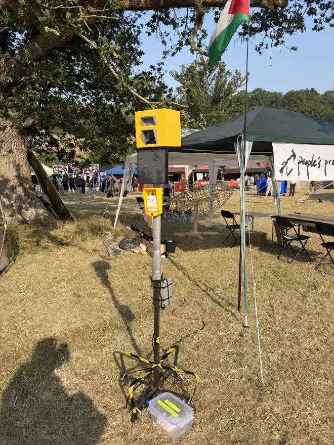 A small yellow speed camera sits on a pole in a field