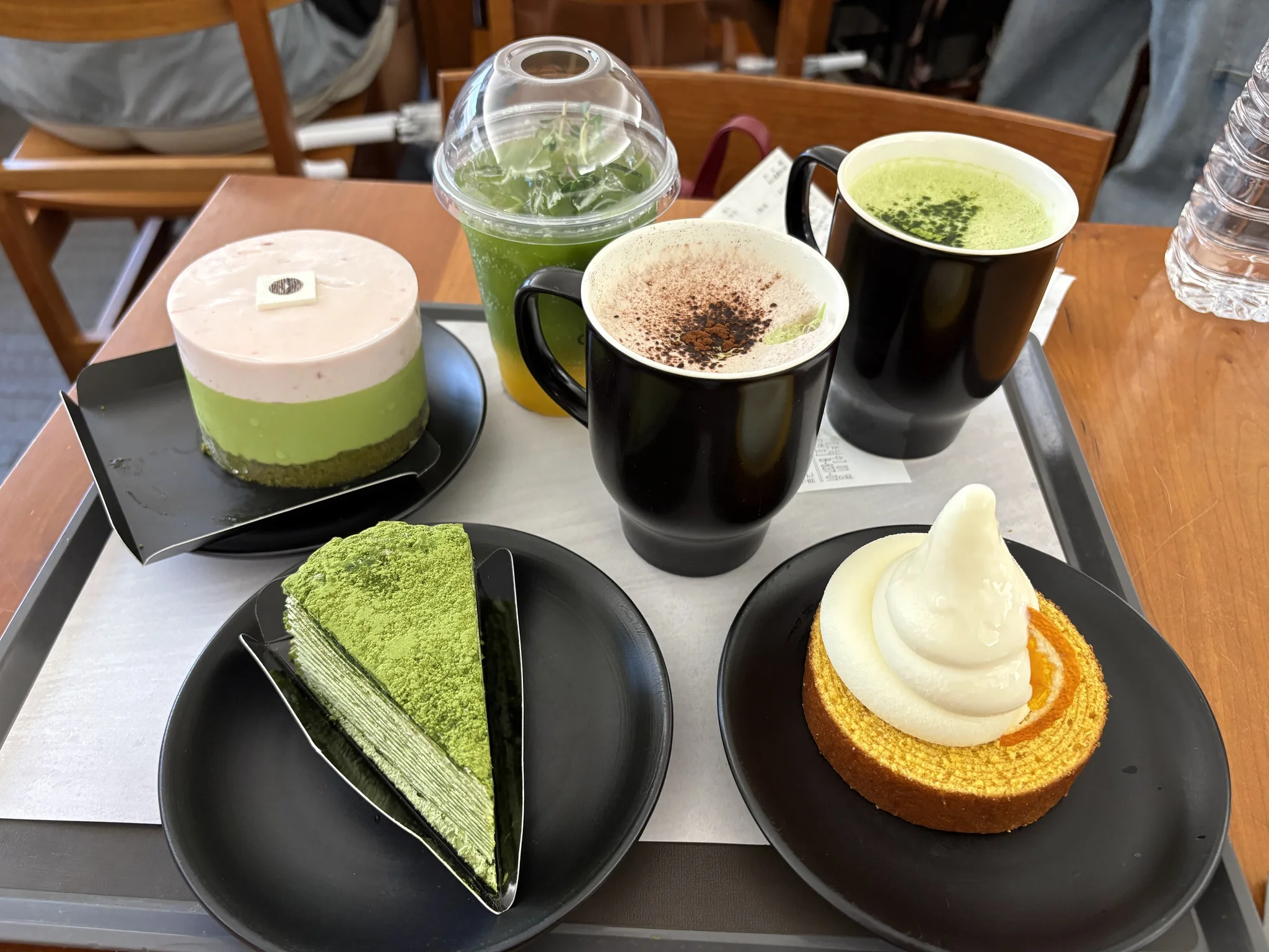 Our lunch at Osulloc Cafe tray displaying three matcha desserts - a layered green tea cake, a mille crepe slice, and a swirled soft-serve tart—alongside two hot beverages and a cold green drink in a café setting
